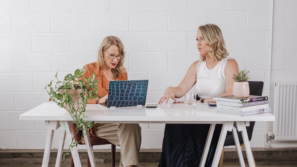 Michelle sits behind a desk on the left with a laptop in front of her. Her client Renata sits on the right. The women are talking.