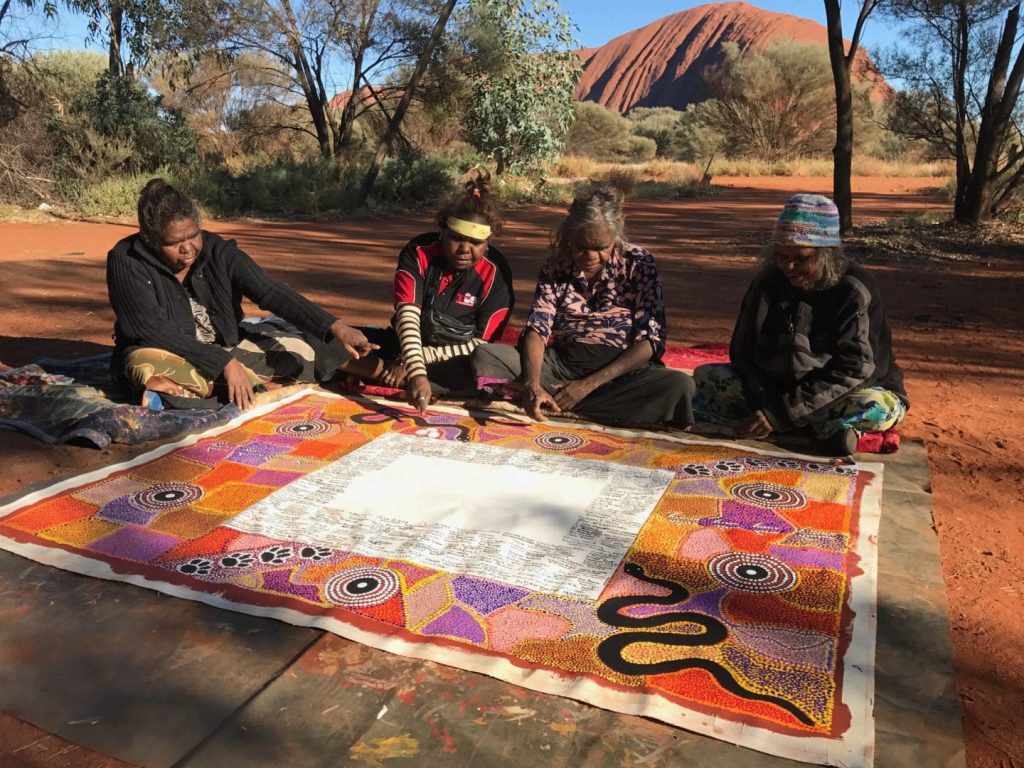 Four Indigenous women sit with the Statement on fabric in front of them, and Uluru behind them.